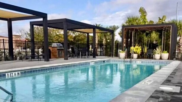 A swimming pool with a clear blue water surface, surrounded by a paved deck and a covered seating area with chairs. - Elegant Home near Disney - pool - free parking (Kissimmee)