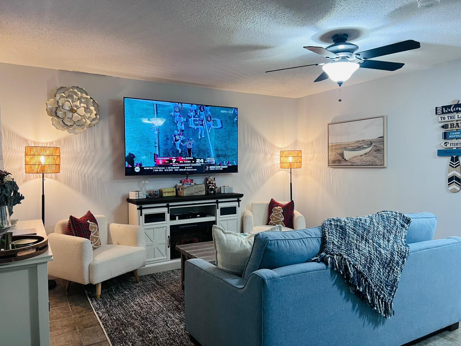 A living room with a flat-screen TV, white and blue furniture, a ceiling fan, and decorative wall art.