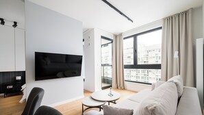 Een moderne woonkamer met een bank, een aan de muur bevestigde televisie, een klein rond tafeltje en stadszicht door grote ramen.