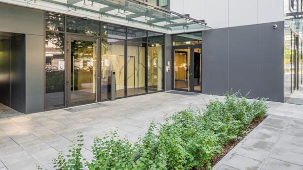 Een modern gebouw met een glazen ingang, een overdekte doorgang en een aangelegde tuin met beplanting.