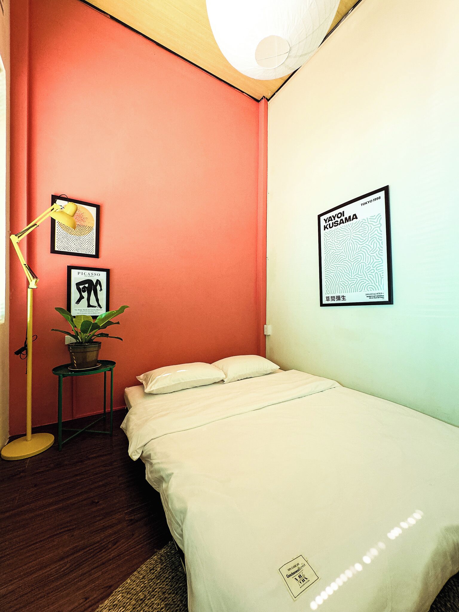 A room with a bed, a desk with a lamp, and a wall with framed artwork.