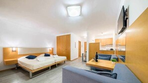Una habitació moderna i ben il·luminada amb un llit, un sofà, una taula petita i una televisió penjada a la paret.