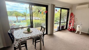Amplia habitación con un gran ventanal que ofrece vistas a una playa y palmeras. Hay una mesa con mantel floral y dos sillas.
