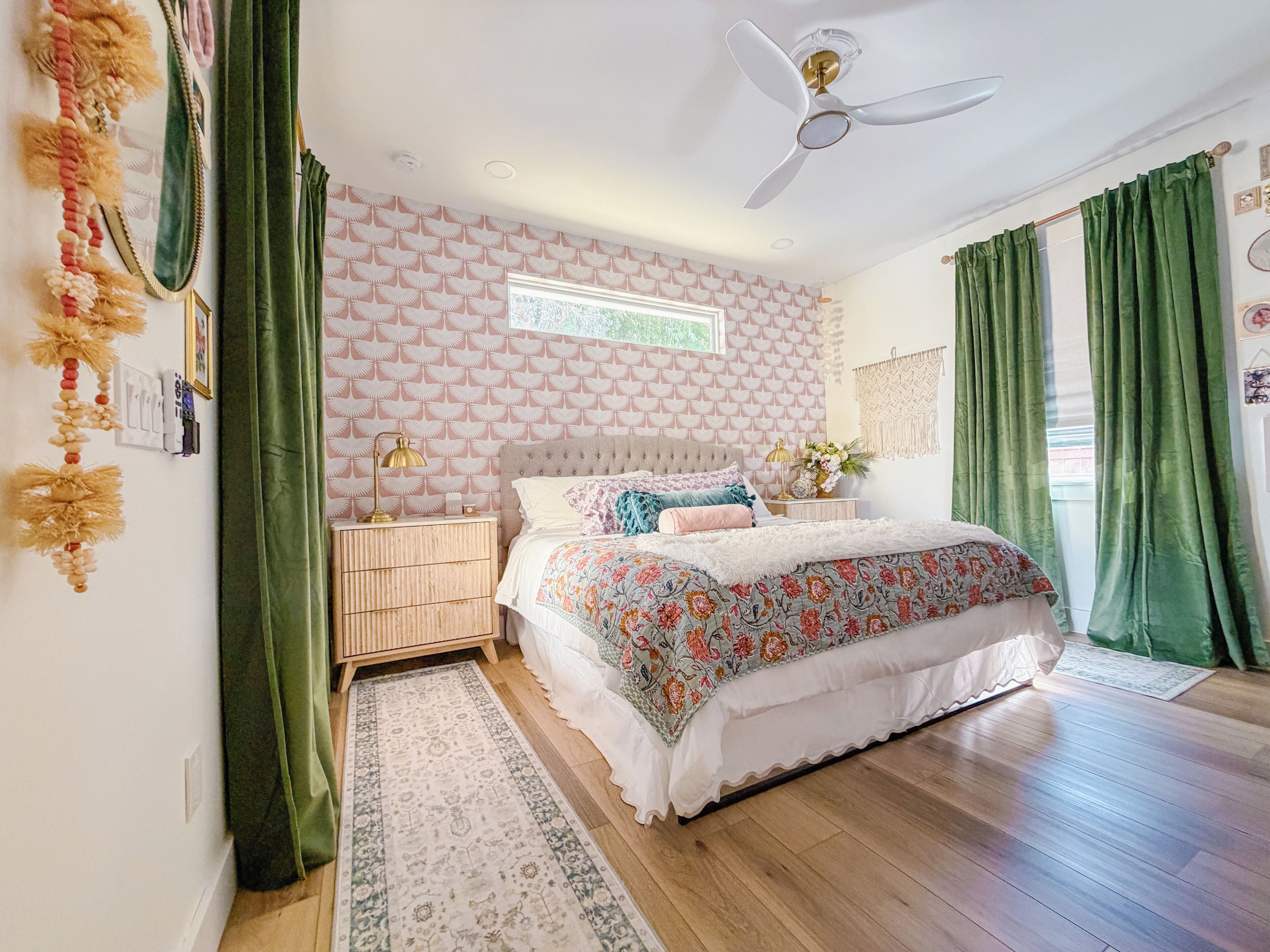 A bedroom with a tufted headboard, a floral bedspread, a standing lamp, and a large window with green curtains.
