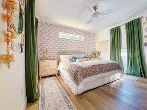 Une chambre à coucher avec une tête de lit capitonnée, une couvre-lit à motifs floraux, un lampadaire et une grande fenêtre ornée de rideaux verts.