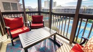Ein Balkon mit zwei roten Sitzgelegenheiten und einem Holztisch, mit Blick auf einen Pool und Gebäude.