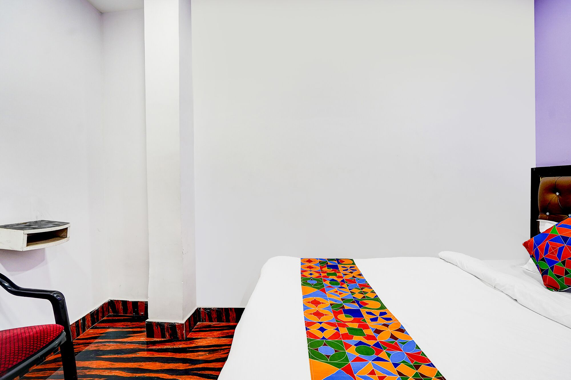 A hotel room with a bed, a chair, a small table, and a colorful geometric-patterned rug.