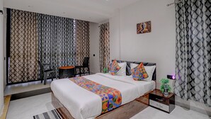 Quarto de hotel moderno com uma cama grande, uma mesinha, duas cadeiras, uma mesa de cabeceira e um vaso de planta.
