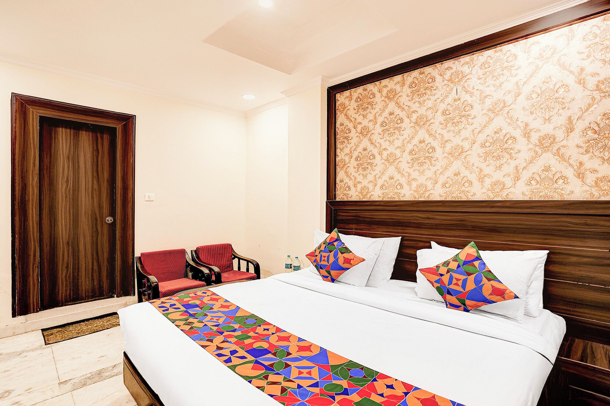 A hotel room with a large bed, two red chairs, a wooden door, and a patterned wallpaper.