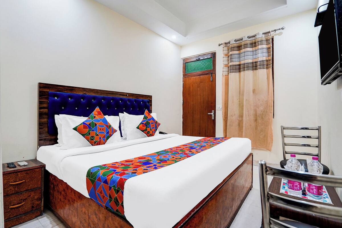 Un dormitorio ordenado con cabecera de madera, una cama con almohadas de diseño geométrico y colores vivos, una colcha blanca y una mesita de noche de madera con un control remoto.