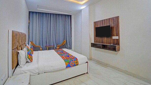A hotel room with a bed, two armchairs, a small table, a TV mounted on a wooden panel, and a window with curtains. - Via Stars (Noida)