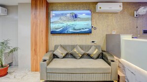 Una zona d'entrada amb un sofà, un televisor fixat a la paret que mostra un paisatge, i un panell de fusta a l'esquerra.