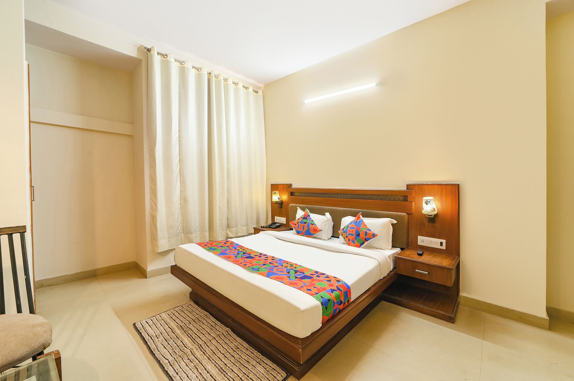 Une chambre d’hôtel comprenant un lit, des tables de chevet, une chaise, une fenêtre avec des rideaux et une lampe fixée au mur.