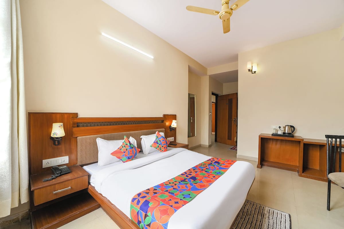A hotel room with a large bed, a wooden headboard, a nightstand with a phone, a desk with a kettle, and a chair.
