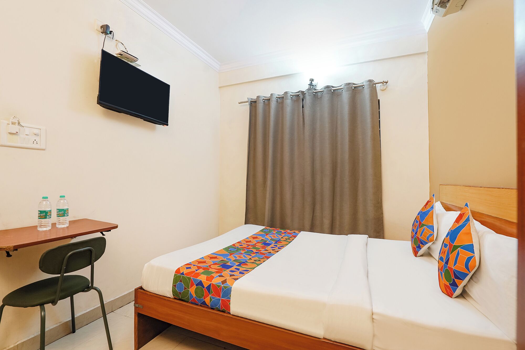 Habitación de hotel con una cama, un escritorio con dos botellas de agua, una silla, un televisor montado en la pared y una cortina.