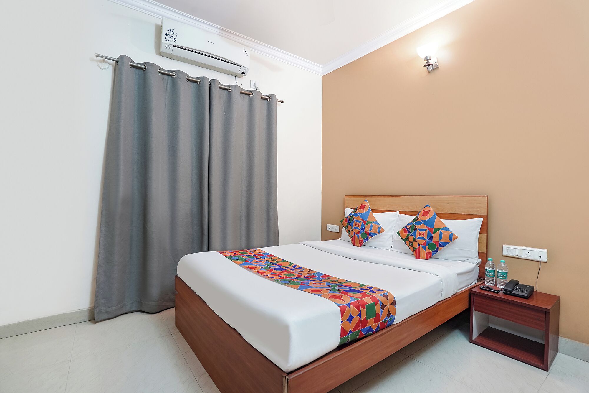 A hotel room with a bed, a nightstand, an air conditioning unit, and a wall-mounted light.