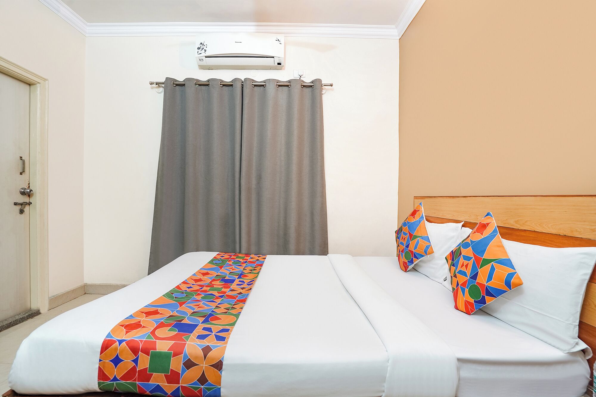 A hotel room with a bed, a headboard, a window with curtains, and an air conditioning unit.