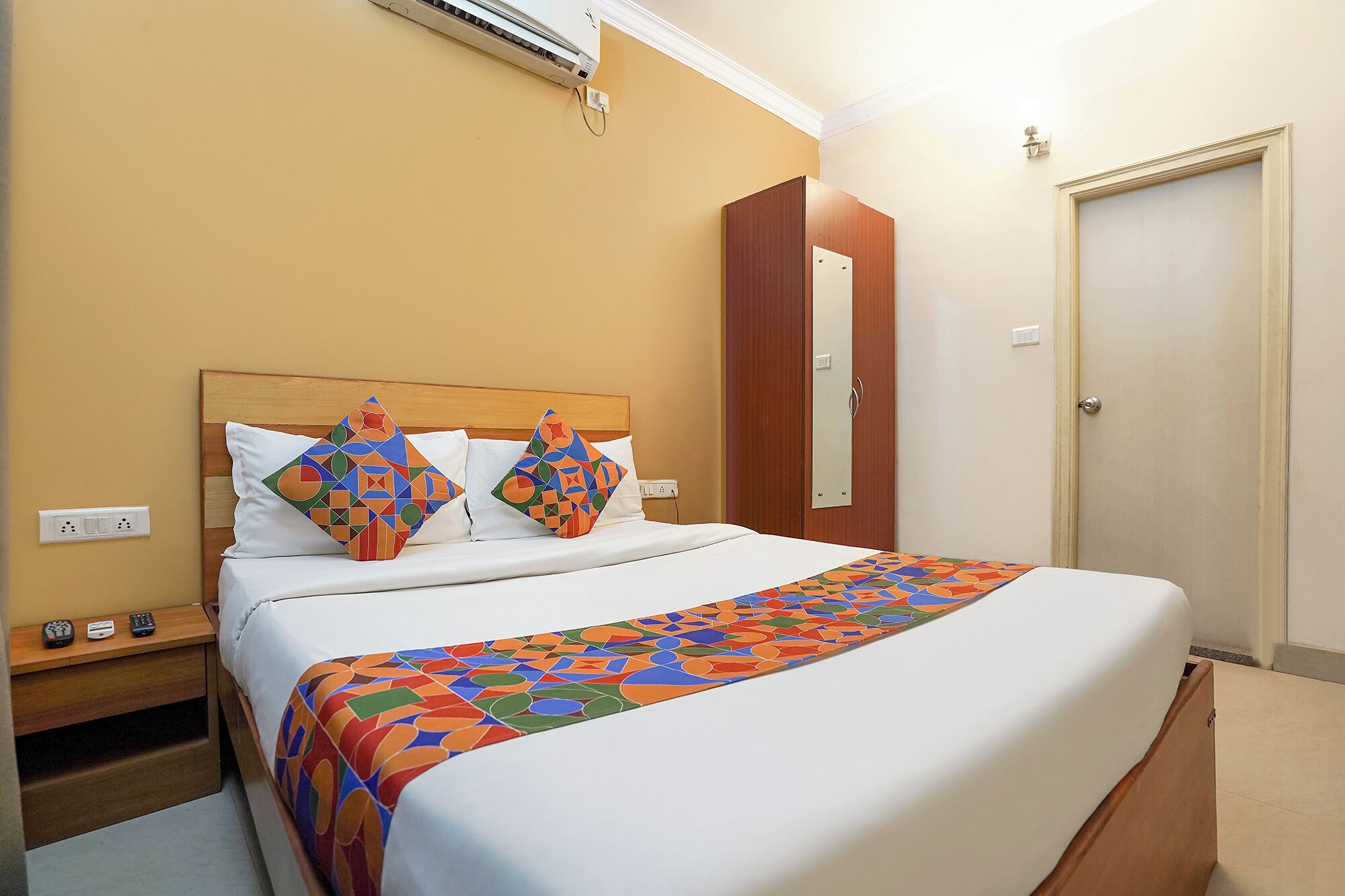 A hotel room with a bed, a nightstand, a bedside table, a wall-mounted air conditioner, and a door.