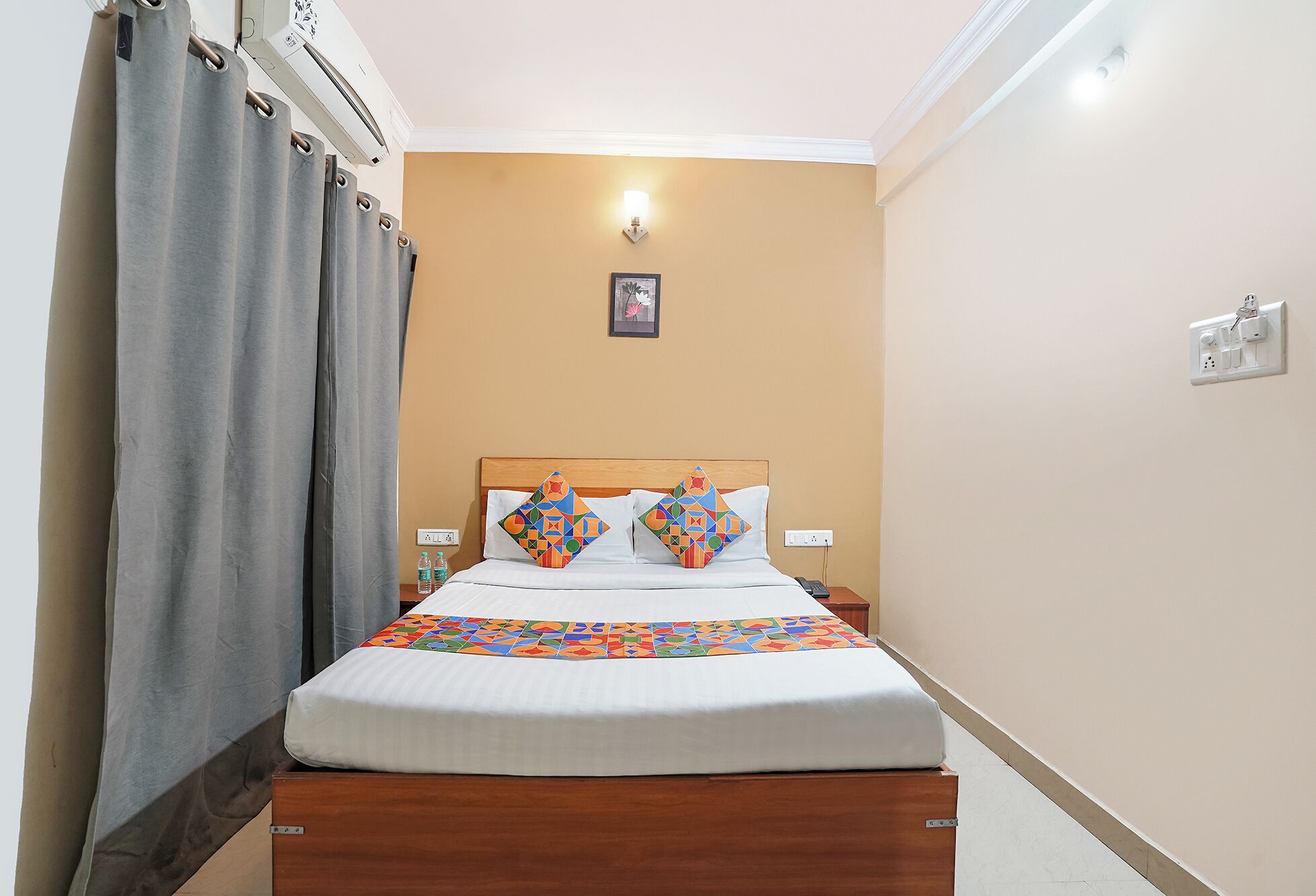 Una habitación de hotel con una cama, cortinas, una mesita de noche y una lámpara de pared, además de un cuadro en la pared.
