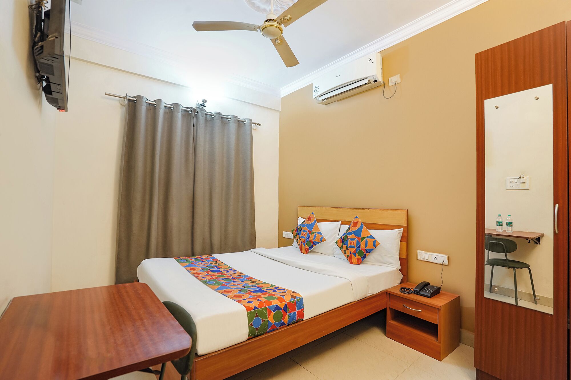 A hotel room with a bed, a desk, a chair, a telephone, a mirror, and a ceiling fan.