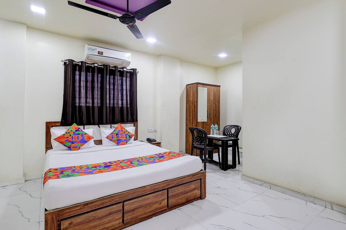 A bedroom with a bed, an air conditioner, a ceiling fan, a wardrobe, a table with chairs, and a mirror.