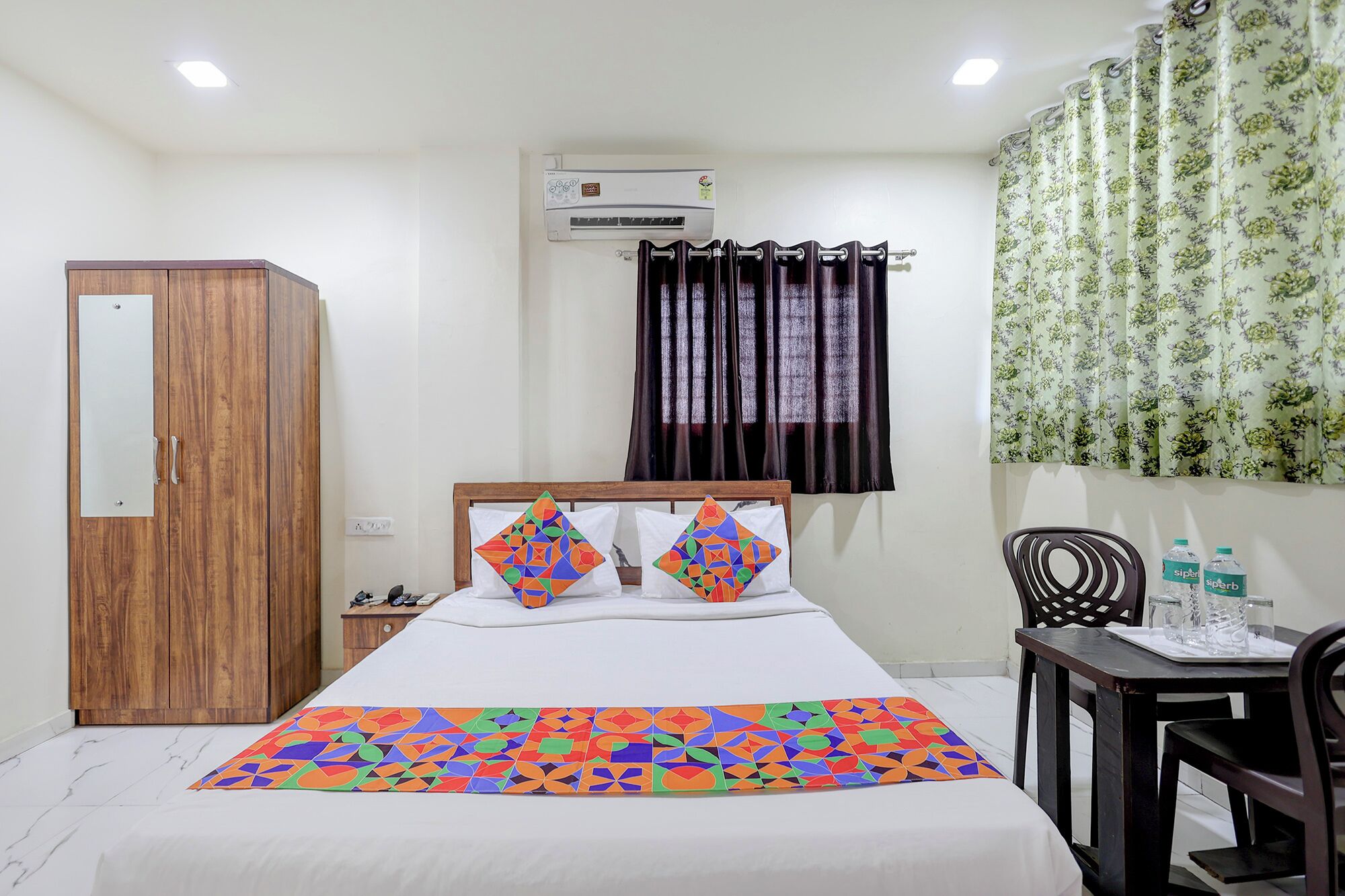 A hotel room with a bed, a wardrobe, an air conditioning unit, a window with dark curtains, and a floral-patterned curtain.
