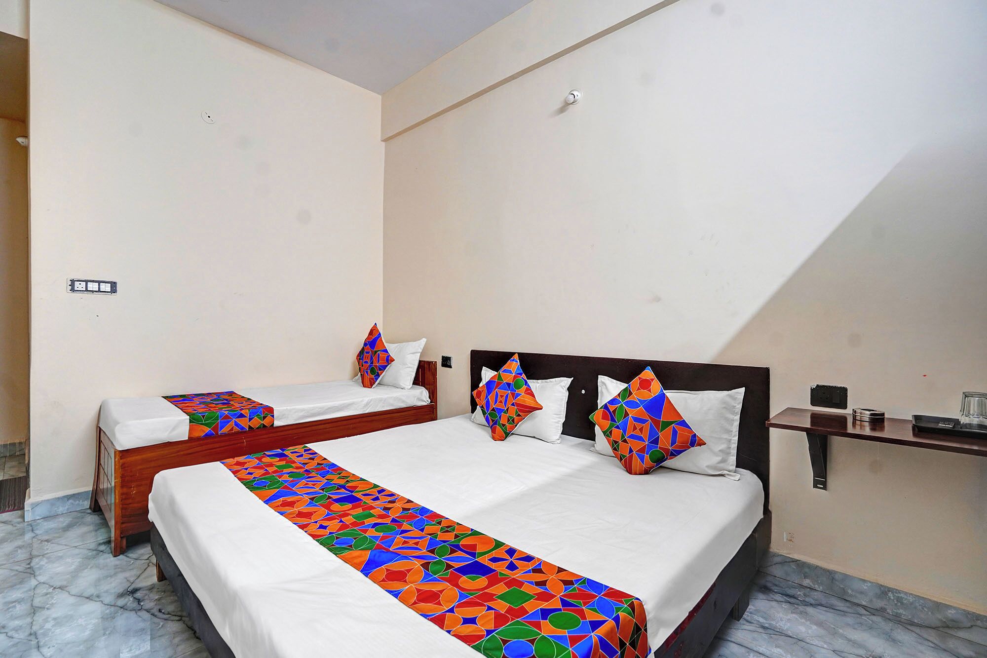 A hotel room with two beds, each with a colorful geometric-patterned bedspread and matching pillows. The room has a simple layout with a wooden headboard and a small table with a glass on it.