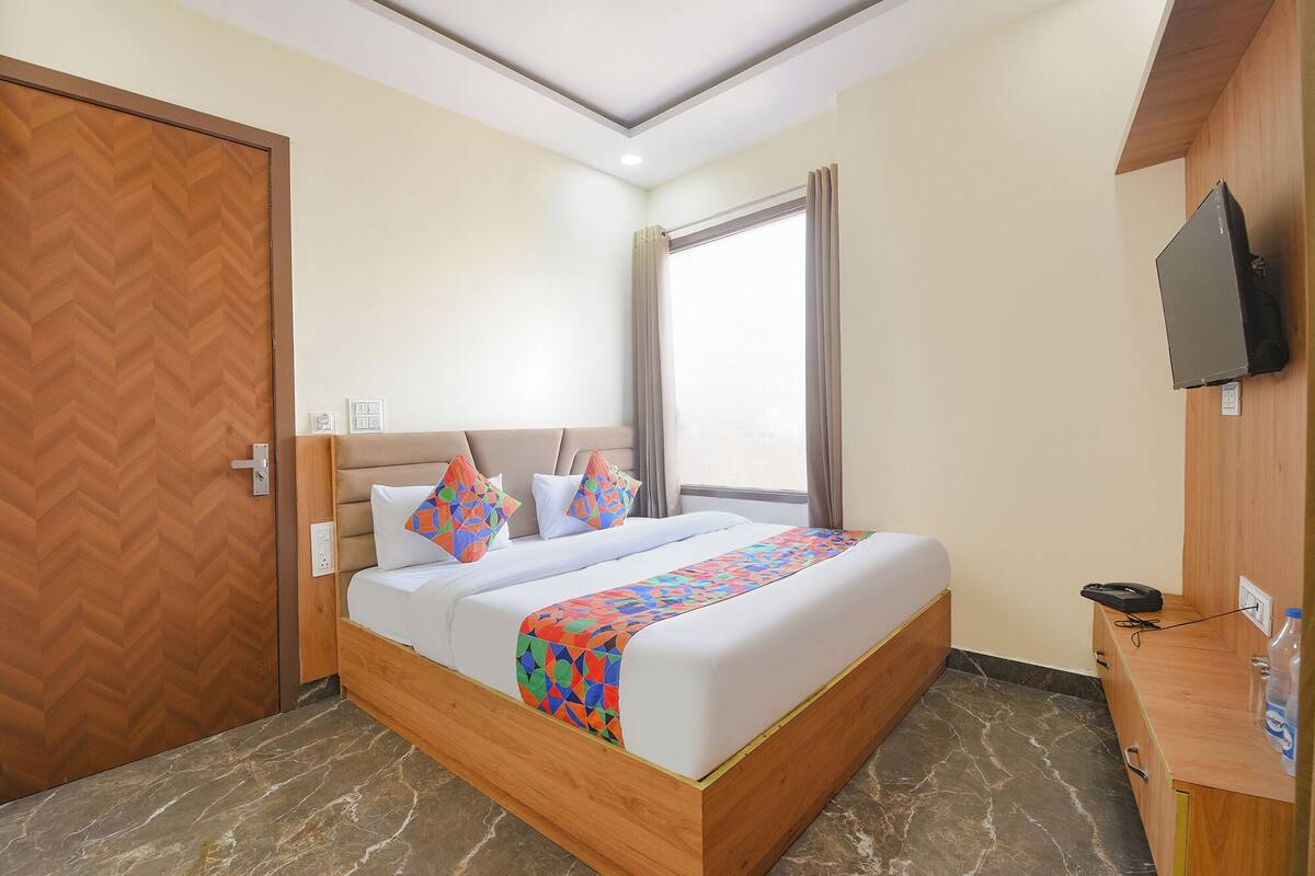A hotel room with a bed, a TV mounted on the wall, a wooden desk, and a window with curtains.