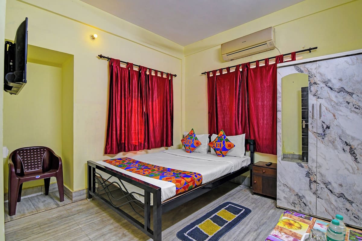 Une chambre d’hôtel avec un lit, des rideaux rouges, une télévision, un climatiseur, un meuble en marbre et une petite table avec une chaise.