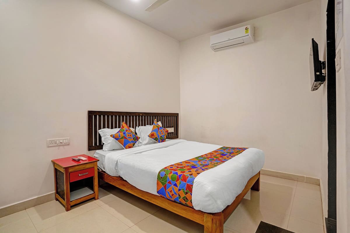 Una cama bien hecha con un edredón de patrón geométrico colorido, un cabecero de madera, una mesita de noche pequeña con tapa roja y una pared blanca con un equipo de aire acondicionado.