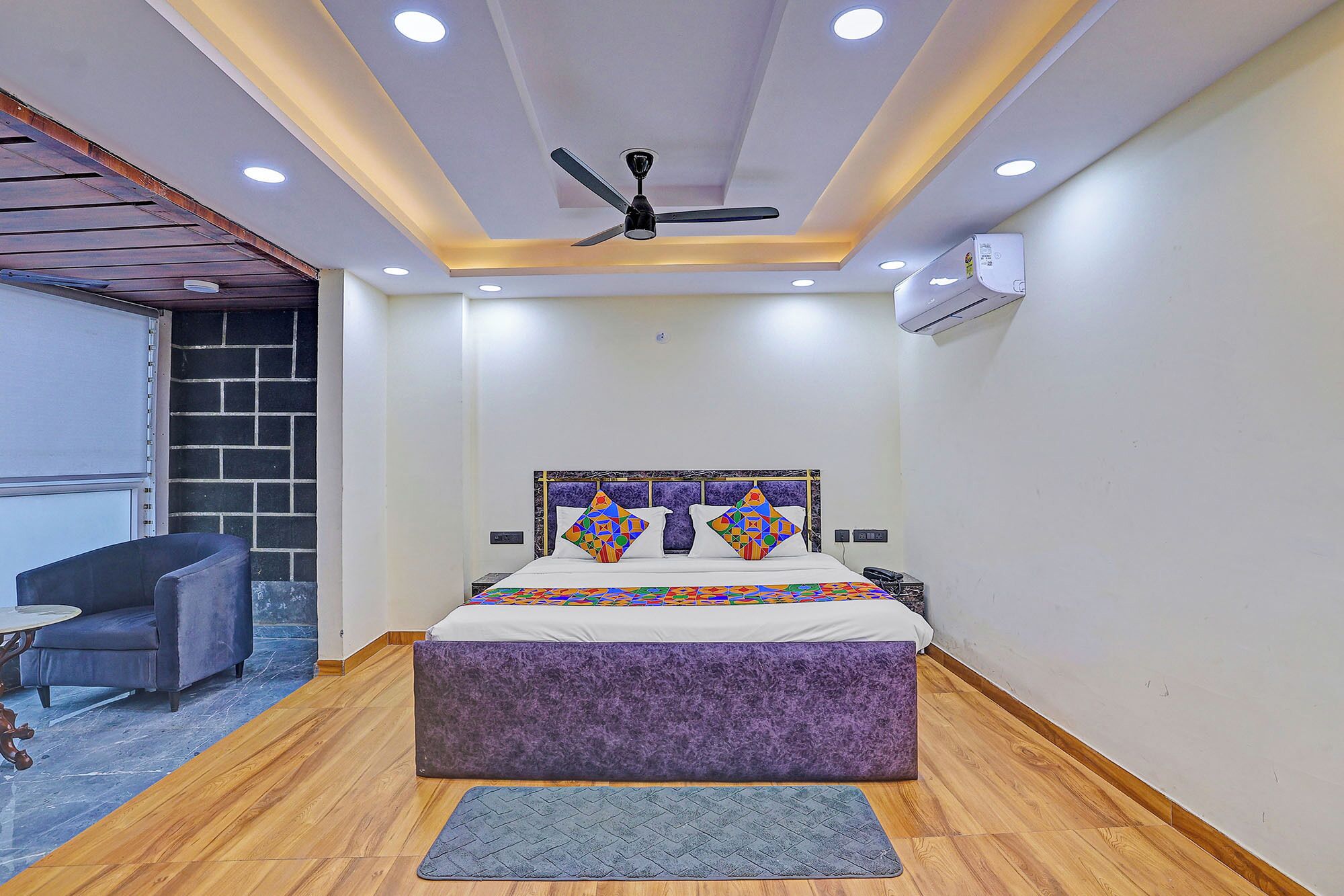 A bedroom with a bed, a ceiling fan, an air conditioning unit, and a small seating area with a chair and table.