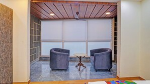 A room with a wooden ceiling, two grey armchairs, a small round table, and a large window with blinds. - FabHotel Havenora - Nr. Maharani Bagh (New Delhi)