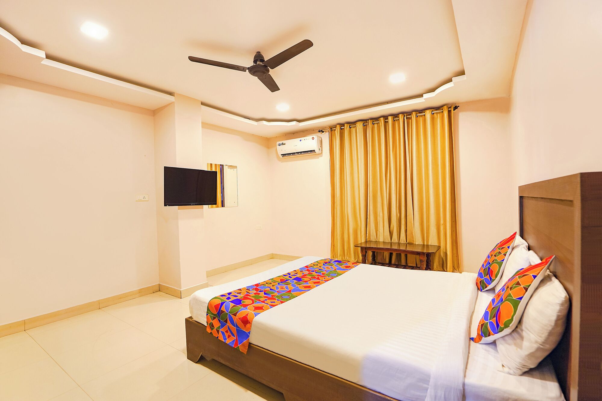 Una habitación de hotel con cama, ventilador de techo, equipo de aire acondicionado, televisor montado en la pared y ventana con cortinas.