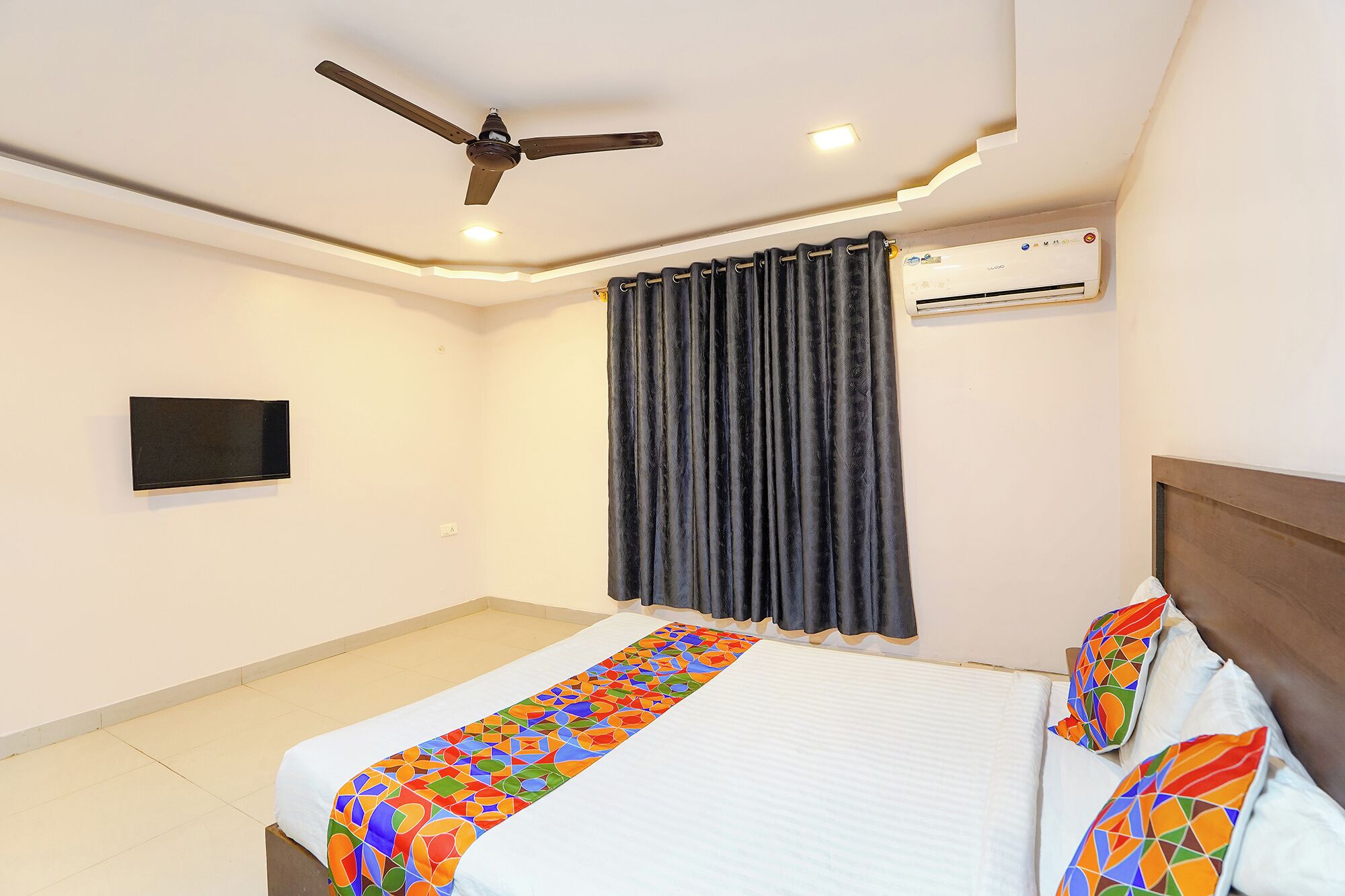 A hotel room with a bed, a ceiling fan, an air conditioning unit, a TV, and a curtain.