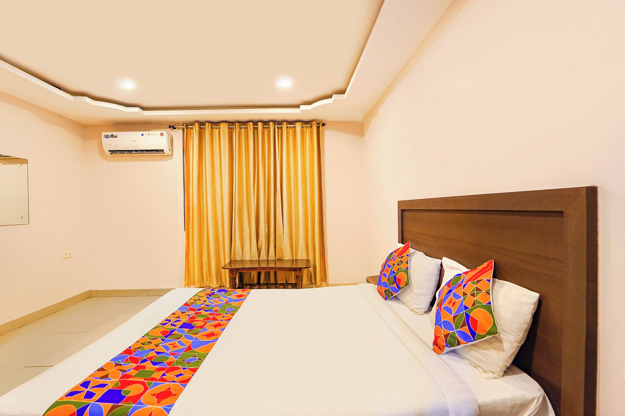 A hotel room with a large bed, a wooden headboard, and a colorful runner. There is a small table and a chair in the corner, and an air conditioning unit on the wall.