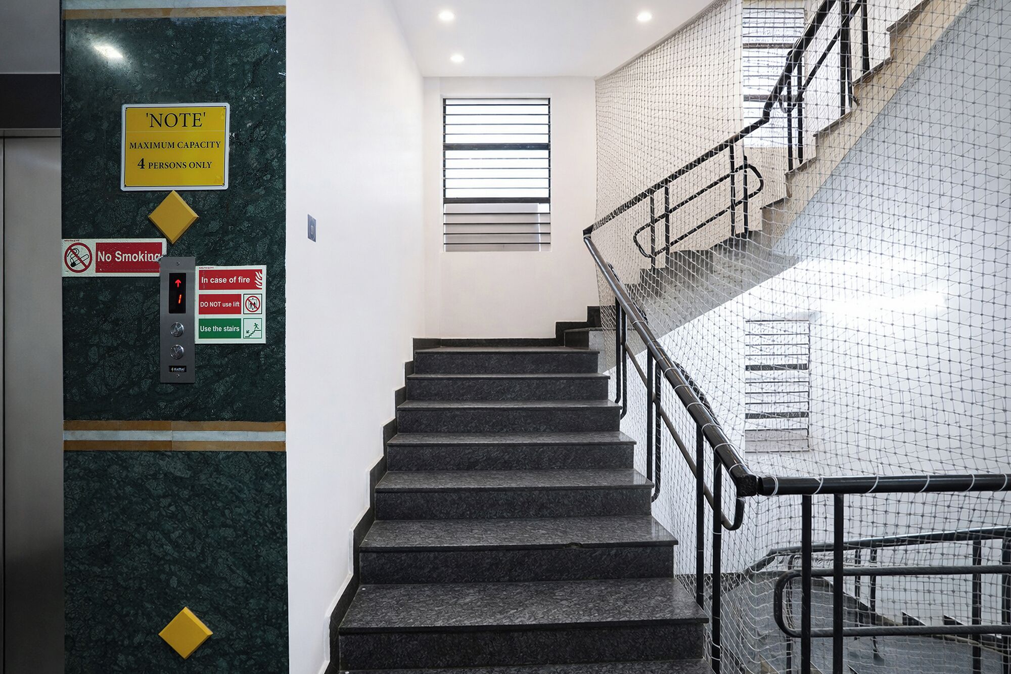 A staircase with a metal railing, a 'No Smoking' sign, a 'Maximum Capacity 4 Persons Only' sign, and a fire safety instruction sign.