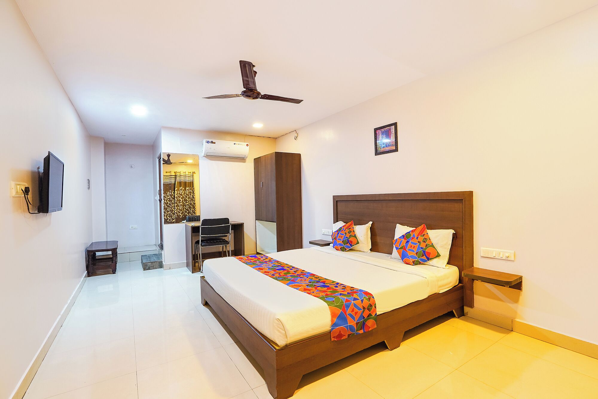 A hotel room with a bed, a desk, a chair, a TV, and an air conditioning unit.