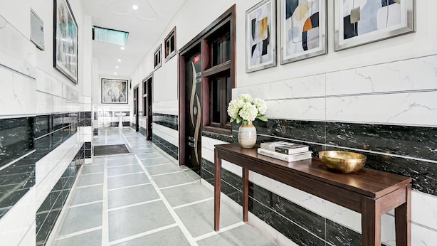 A hallway with a marble floor, framed artwork on the walls, and a wooden table with a vase of flowers. - Fabhotel Happy Stay I (Noida)