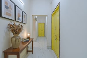 A hallway with a wooden console table, a vase with dried flowers, framed artwork on the wall, and a bright yellow door at the end. - Fabhotel Tulip (Kolkata)
