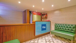 A reception area with a wooden reception desk, a blue 'Via' sign, a green sofa, and a patterned floor. - Via Woodlands (Mumbai)