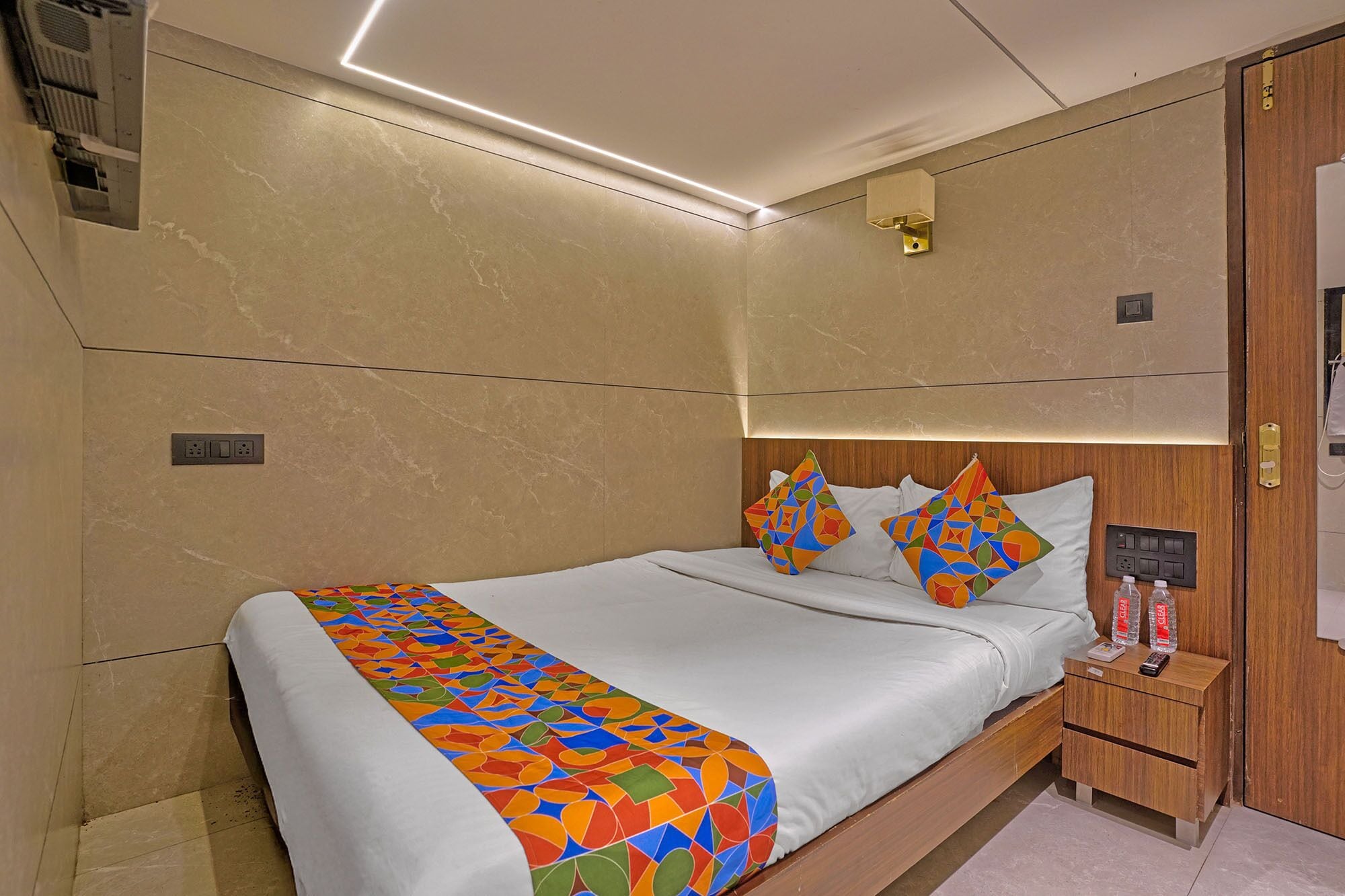 A modern hotel room with a neatly made bed, a bedside table with a remote control, and a wall-mounted light fixture.