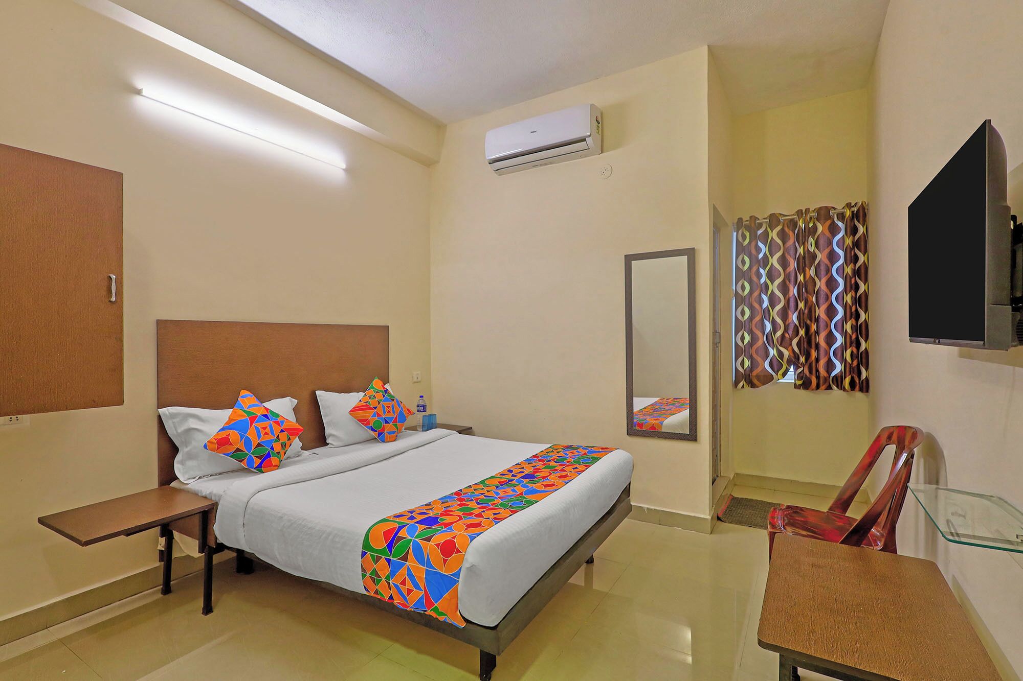 A hotel room with a bed, a desk, a chair, an air conditioner, a mirror, and a window with curtains.