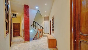 A hallway with a marble floor, a wooden door, a staircase with a red brick pillar, and a white door at the end. - Via Crown DX (New Delhi)
