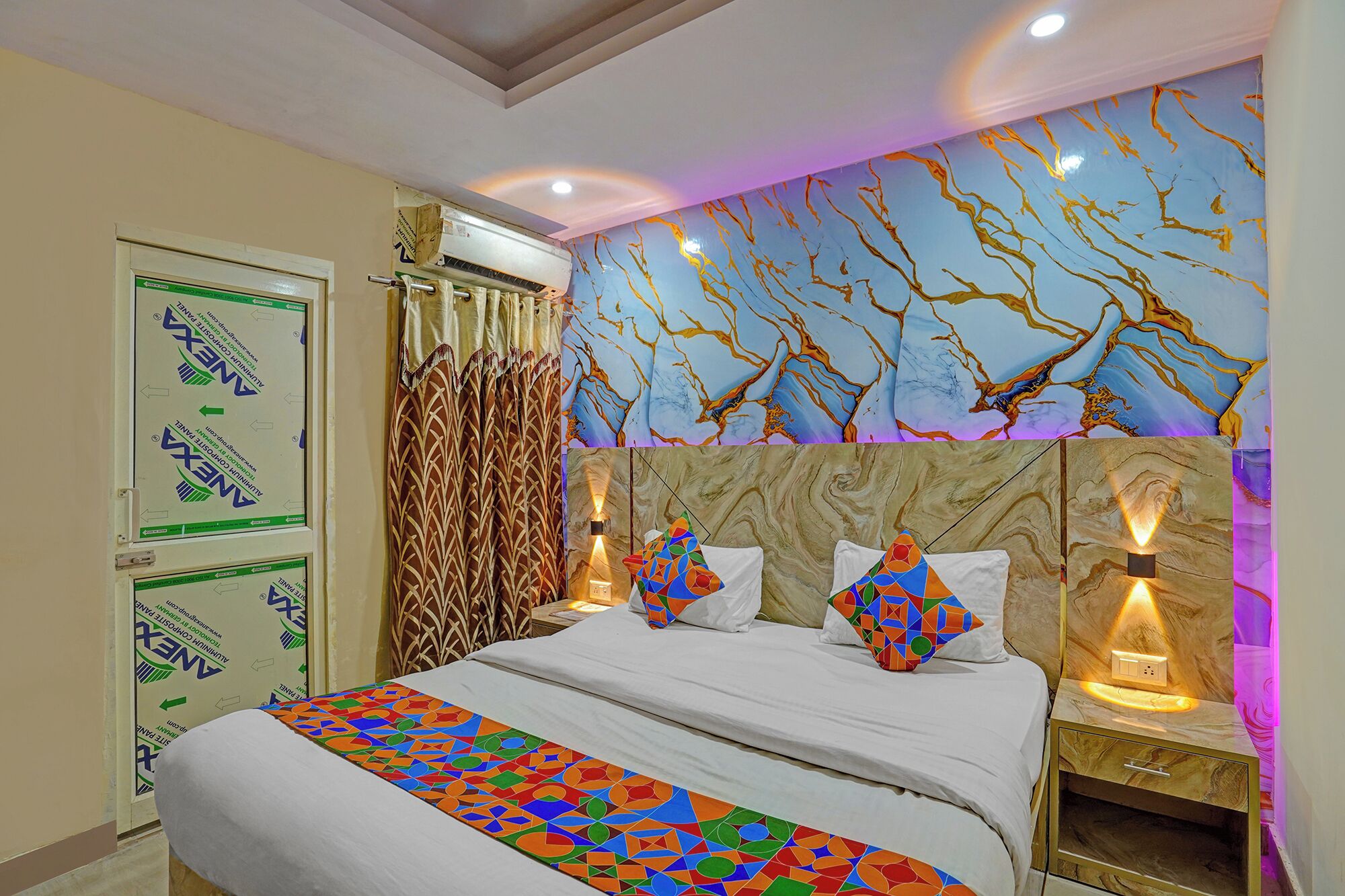 A hotel room with a bed, a nightstand, a wall with a tree branch mural, and a window with curtains.