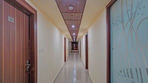 A long, narrow hallway with wooden doors on one side and a glass door with a tree design on the other. The ceiling has recessed lighting and a smoke detector. - Fabhotel Mj International (Lucknow)