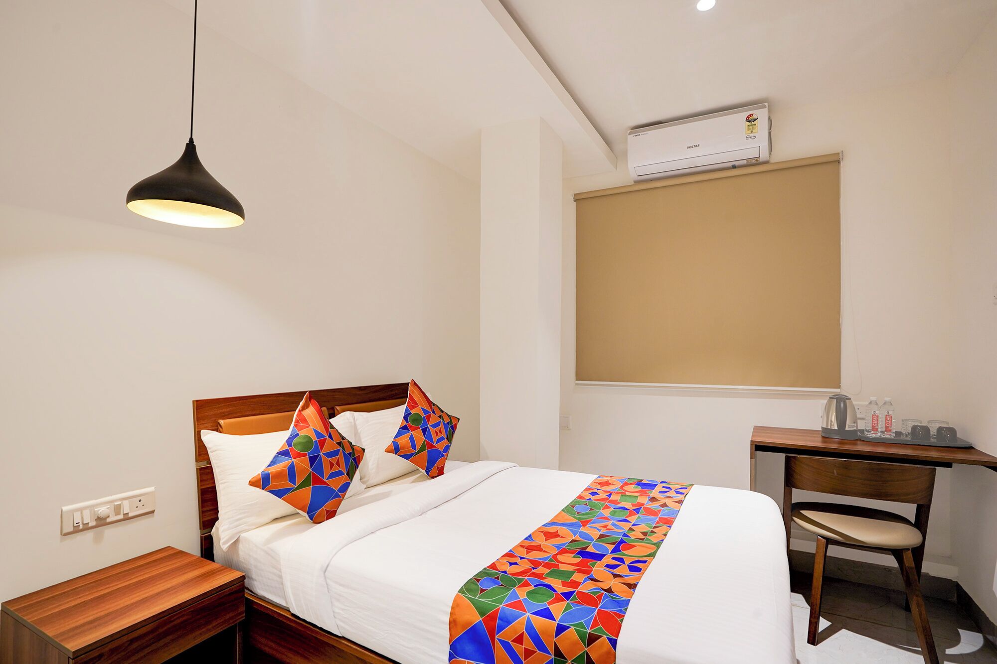 A hotel room with a bed, a desk, a chair, an air conditioner, and a hanging light fixture.