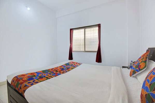 A neatly made bed with a colorful runner, a window with curtains, and a plain white wall. - Via Royal Stay I (Pune)