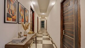 A hallway with a checkered floor, framed artwork on the walls, a wooden door, and a console table with books and a vase. - Fabhotel Satya (Noida)