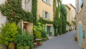 Eine schmale Straße, gesäumt von Gebäuden, die in Grün bewachsen sind, mit Kübelpflanzen und einem kleinen Tisch mit Stühlen auf dem Bürgersteig.