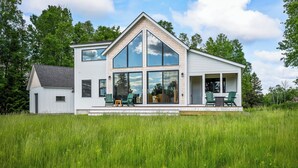 A two-story house with a large glass front and a porch with chairs. - The Birches on Marbles Way | Snowmobile Access | Waterfront Access on Rangeley Lake (Rangeley)
